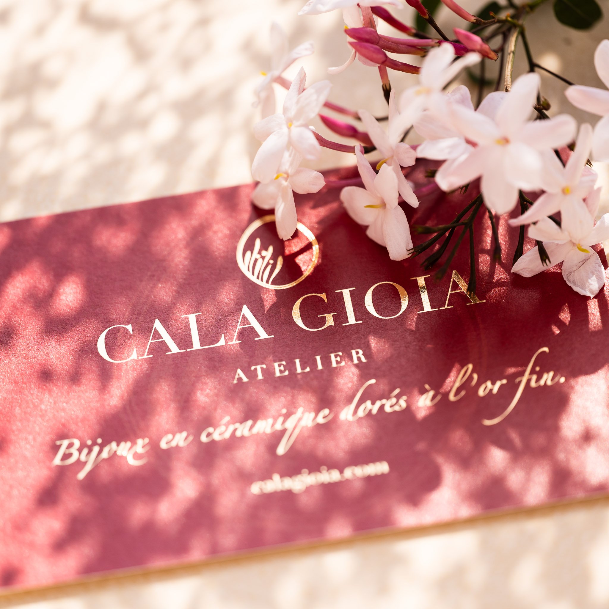 Carte d'authenticité texturée Cala Gioia posée délicatement parmi des fleurs de jasmin fraîches. Une invitation à faire confiance à l'artisan pour la sélection d'un bijou mystère en céramique et or fin.