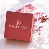 Écrin à bijoux terracotta au logo doré Cala Gioia, délicatement posé à côté de fleurs de jasmin odorantes. Illustration du concept "Le Cadeau Parfait", une expérience mystère où l'artisan sélectionne avec intuition votre futur bijou en céramique et or fin.