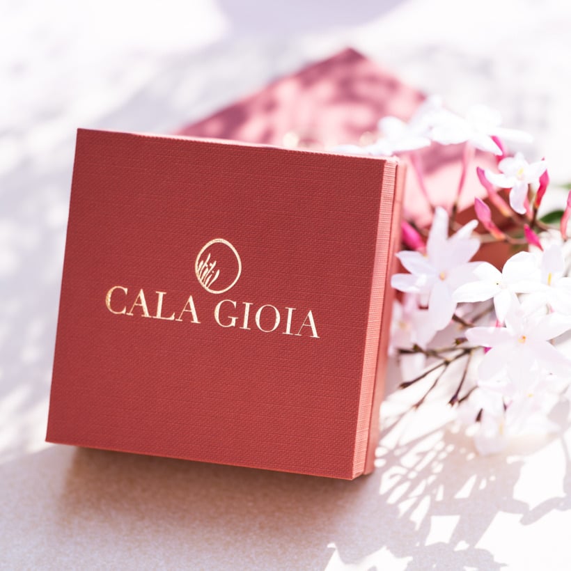 Écrin à bijoux terracotta au logo doré Cala Gioia, délicatement posé à côté de fleurs de jasmin odorantes. Illustration du concept "Le Cadeau Parfait", une expérience mystère où l'artisan sélectionne avec intuition votre futur bijou en céramique et or fin.