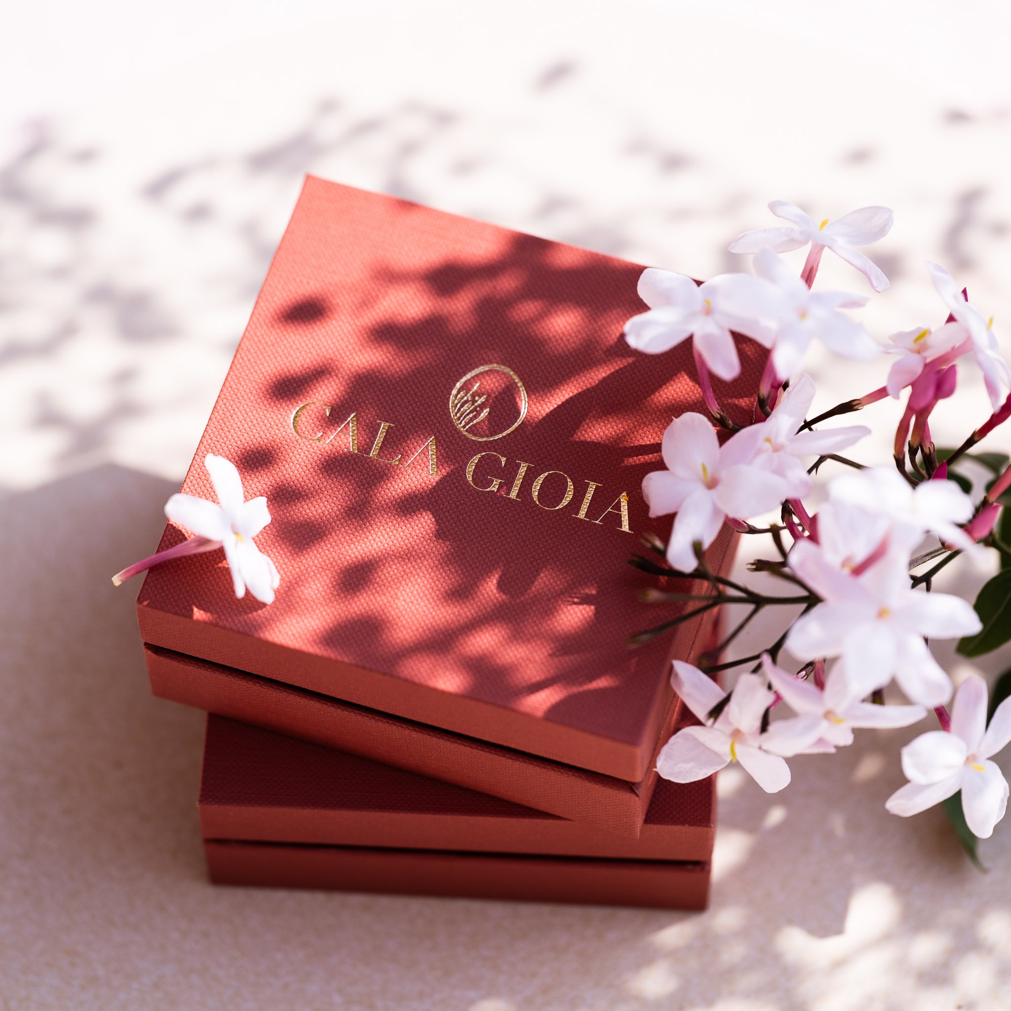 Deux luxueux écrins terracotta Cala Gioia superposés près de quelques fleurs de jasmin. Le moment de la révélation du paquet précieux contenant votre bijou sur-mesure, choisi selon vos nuances préférées.