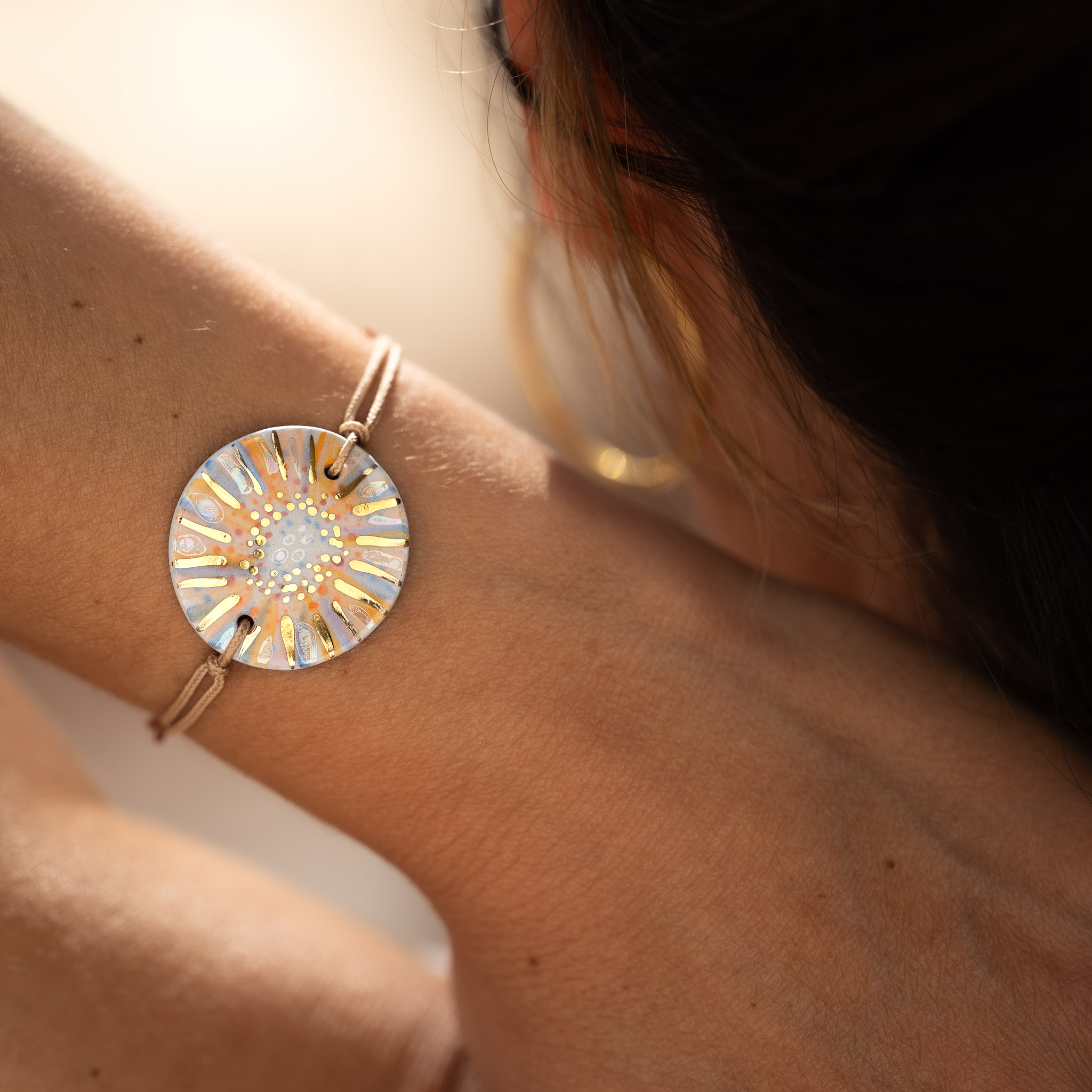 Un bracelet CALA GIOIA aux nuances orange et bleu, née de trois feux successifs, capturant l'éclat de la lumière et les secrets de la terre sur le poignet d'une femme de dos.