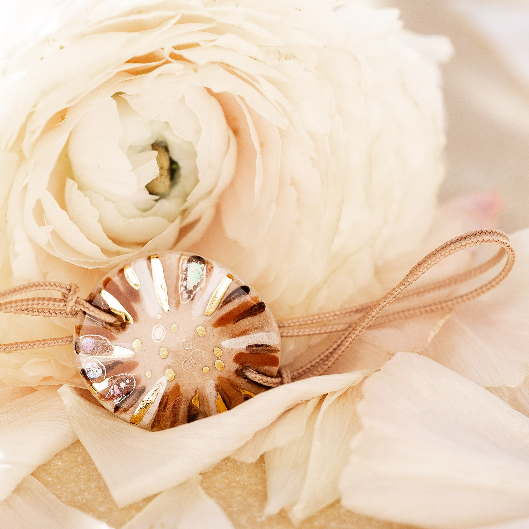 Notre engagement de durabilité : un bijou aux nuances brunes Cala Gioia reposant sur une rose blanche, symbolisant la rencontre entre la force de la terre et la délicatesse du temps.