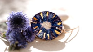 Détail des nuances de cuisson appelées Ombre Marine sur un bracelet bleu en céramique doré à l'or fin Cala Gioia, entouré de fleurs de séné de Provence