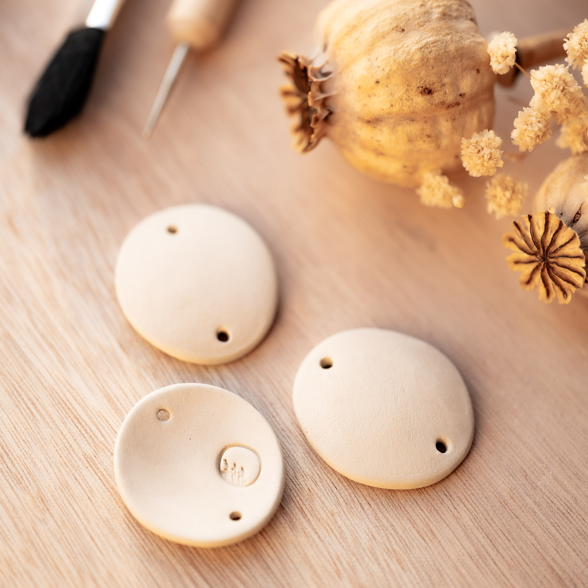 L’éloge de l’imparfait : trois ébauches de bijoux en terre crue reposant auprès de nos outils de modelage et de pavots séchés, célébrant le geste souverain de la main sur la matière brute.