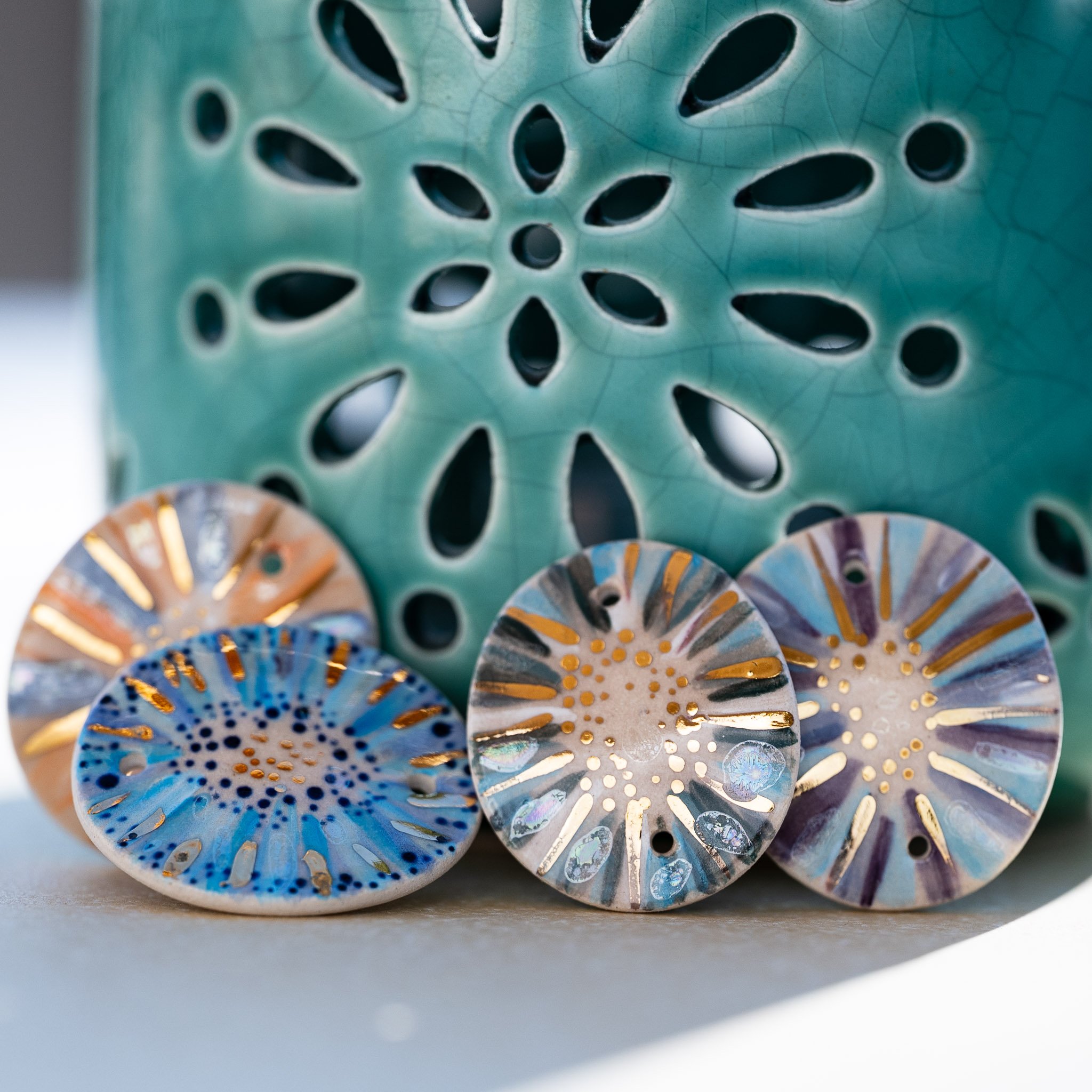 Capturer le soleil et l’eau : quatre médaillons Calagioia aux reflets azurés et violets, dont l’or pur scintille devant un photophore artisanal bleu aux motifs floraux