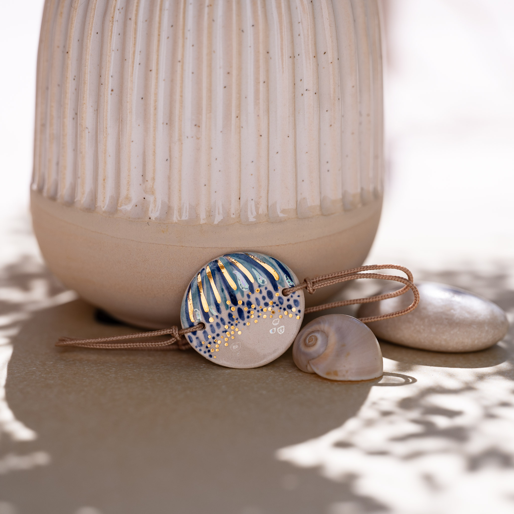 Bracelet Rivage Ensoleillé en faïence bleu marine et or, près d'un vase, d'un galet et d'un œil de Sainte-Lucie - Cala Gioia